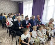 На фотографии изображено мероприятие, посвященное Дню Победы. В актовом зале собрались люди разных возрастов. Многие из них носят георгиевские ленточки. Обстановка торжественная, зрители внимательно слушают выступление, создавая атмосферу патриотизма и уважения к памяти героев.