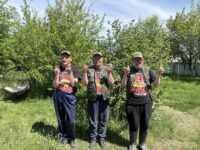 На фотографии изображены три мужчины, стоящие на фоне зелёных деревьев и кустарников. Они одеты в чёрные футболки с надписью «Самый главный рыбак в мире» и изображением, рыбкой. На них также надеты зелёные жилеты. Каждый из них держит в руках рыбку.