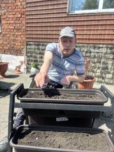На фото человек в инвалидной коляске сидящий на улице,сеет семена в длинные прямоугольные цветочные ящики, установленные на многоярусной подставке. На ящике наклейка с надписью «УКРОП».
