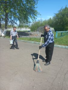 На фото два мужчины на асфальтированной площадке. Один подметает метлой, другой держит лейку в руке