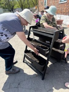На фото — уличная сцена с многоярусной подставкой для растений: в трёх пластиковых ящиках лежит земля. Рядом несколько человек, один сеет семена в ящик, у другого в руках пакетик с семенами. Похоже на посадку или подготовку грядок в контейнерах